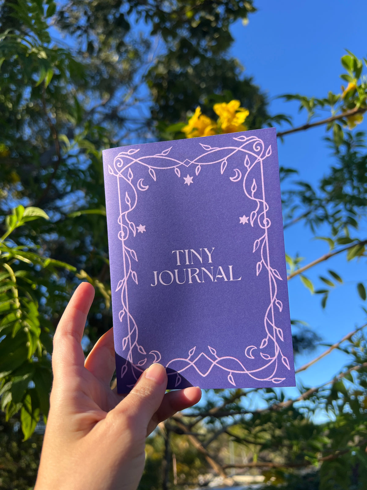 A hand holds a small purple journal with white vine illustrations and the text 'TINY JOURNAL' against a background of green trees and blue sky as a part of the ordinary magic mail club